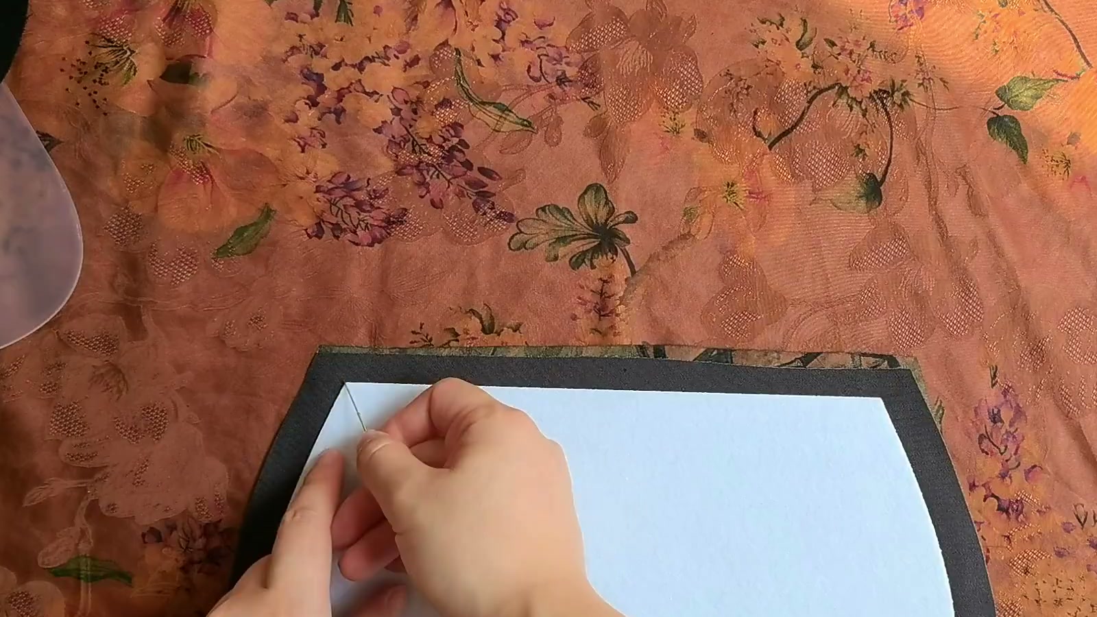 The floral flap and black outer back panel are right sides together while a hand needle sews along their aligned white batting edges